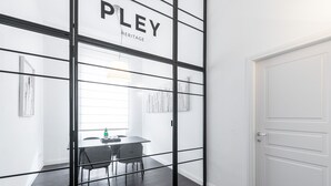 A modern room with a glass partition labeled "PLEY HERITAGE," a white door, and a dining area with a table and chairs.