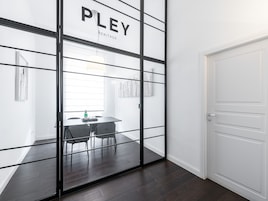 Una habitación moderna con un panel de vidrio que indica “PLEI HERITAGE”, una puerta blanca y una zona de comedor con mesa y sillas.