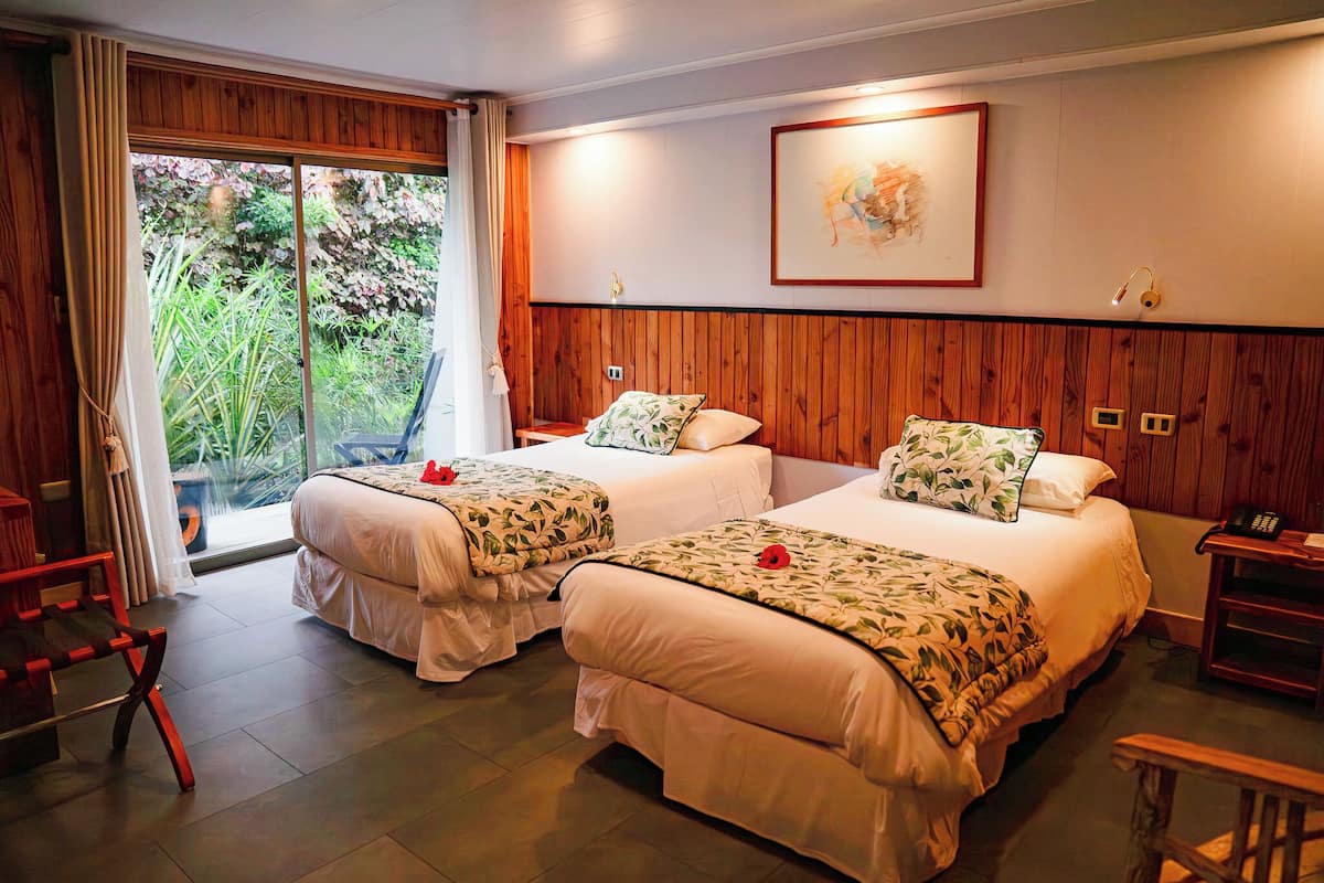 A hotel room with two beds, a sliding glass door to a garden, a framed picture on the wall, and a bedside table with a lamp.