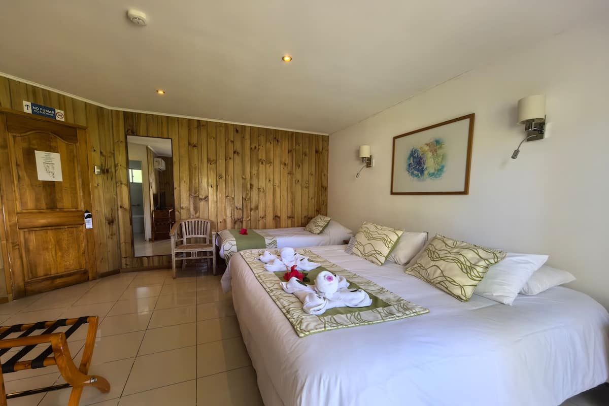A hotel room with two beds, wooden paneling, a chair, a table, and a painting on the wall.