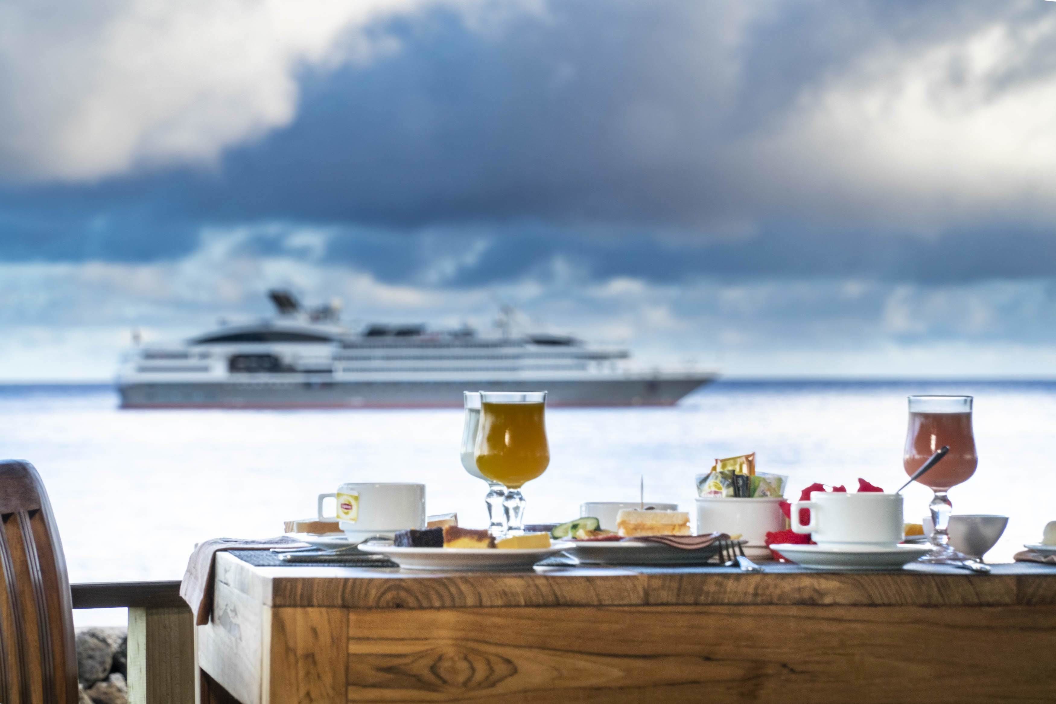 A table set with breakfast items and a glass of tea, overlooking a body of water with a cruise ship in the distance.