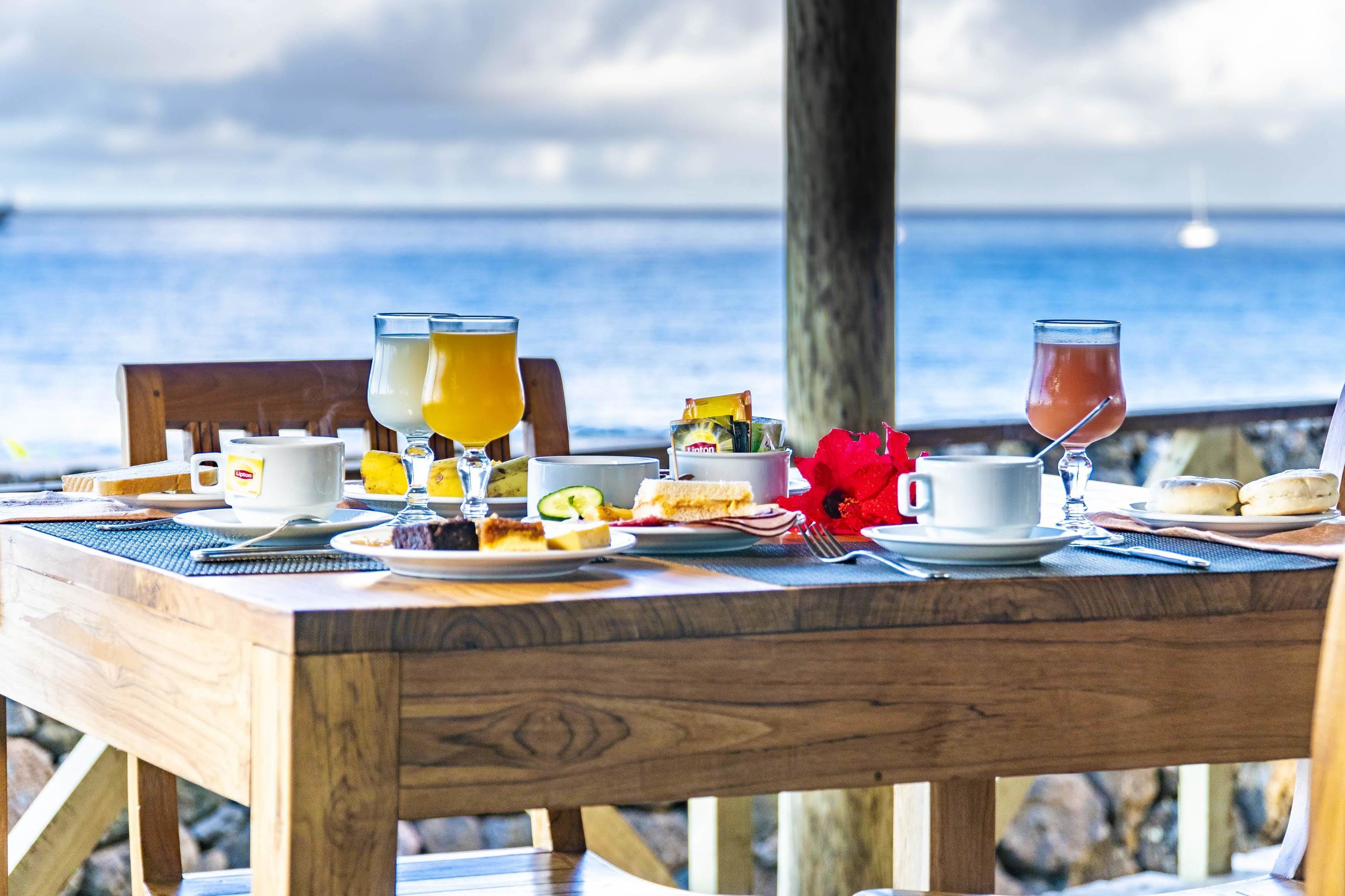 A wooden table set with breakfast items and drinks, overlooking the ocean.