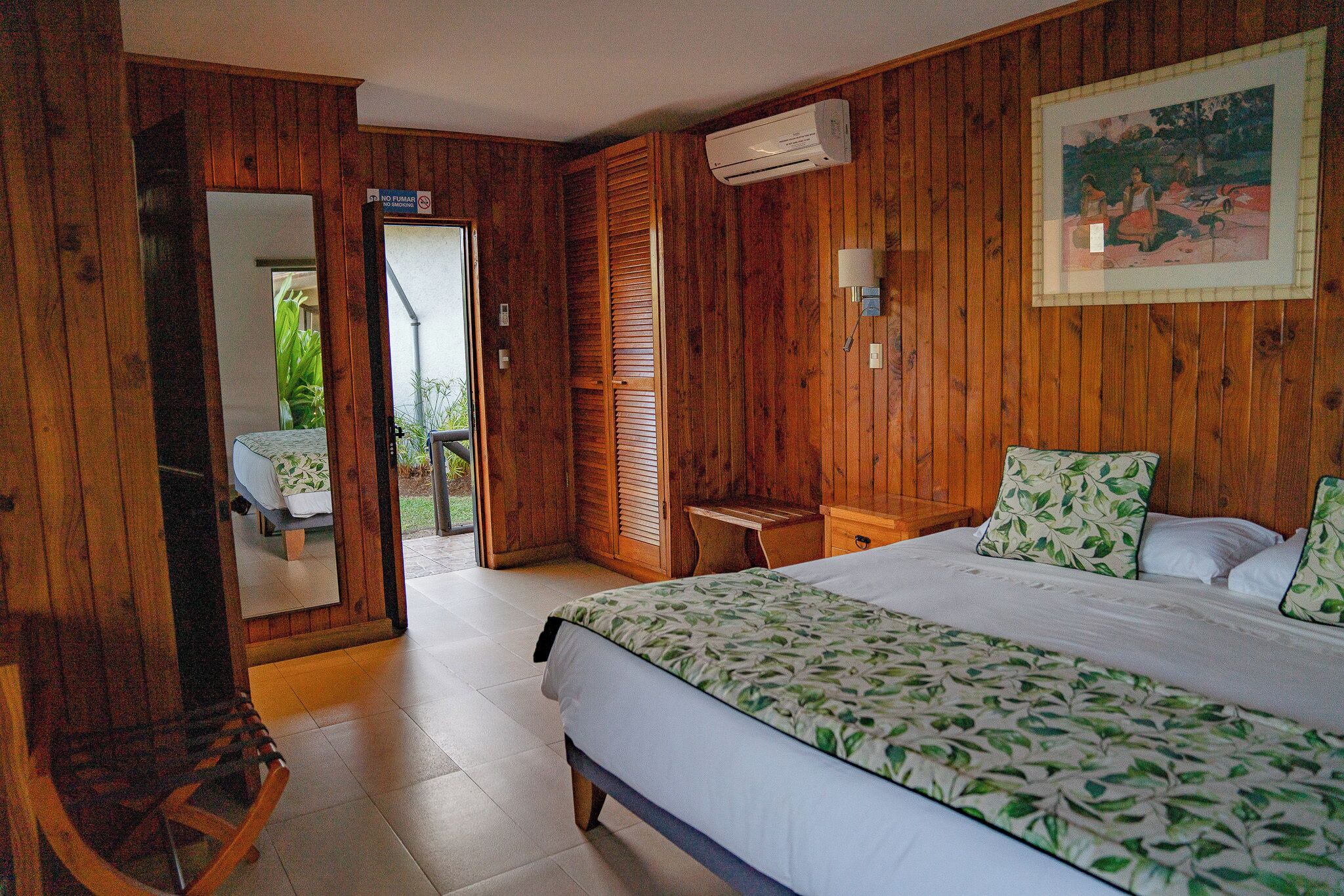 A wooden-paneled room with a bed, a chair, a small table, and a door leading outside.