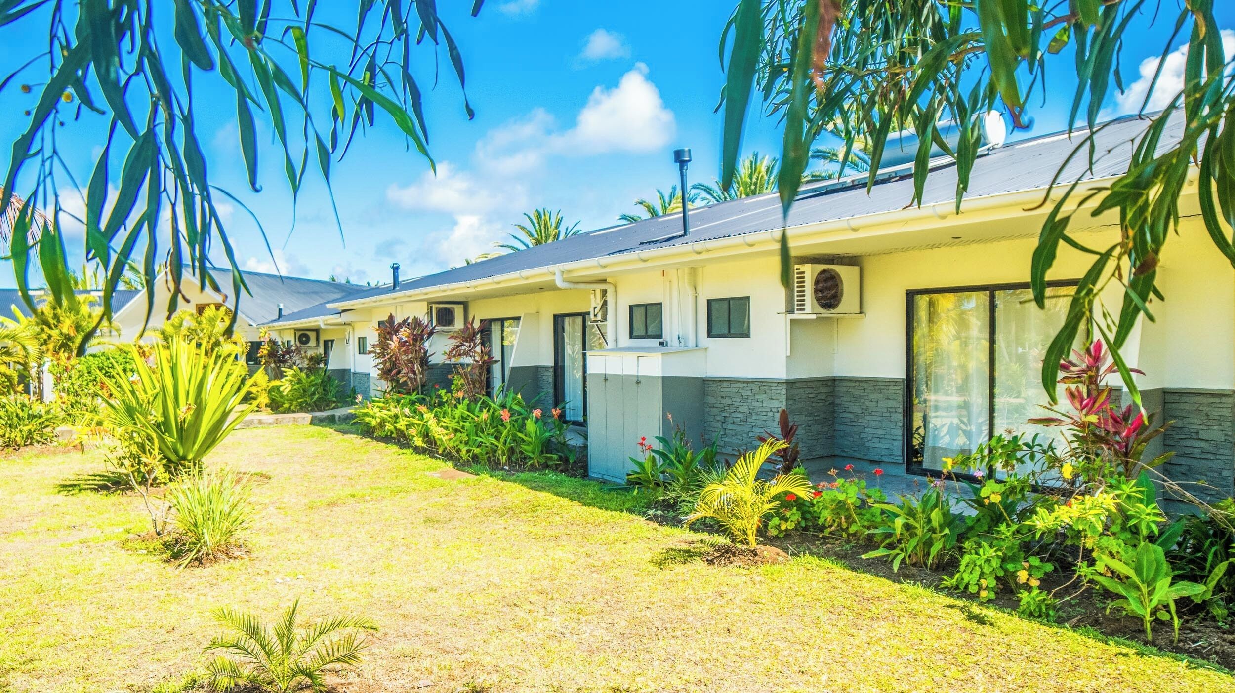 A row of modern, single-story buildings with large windows and air conditioning units, surrounded by well-maintained gardens and tropical plants.