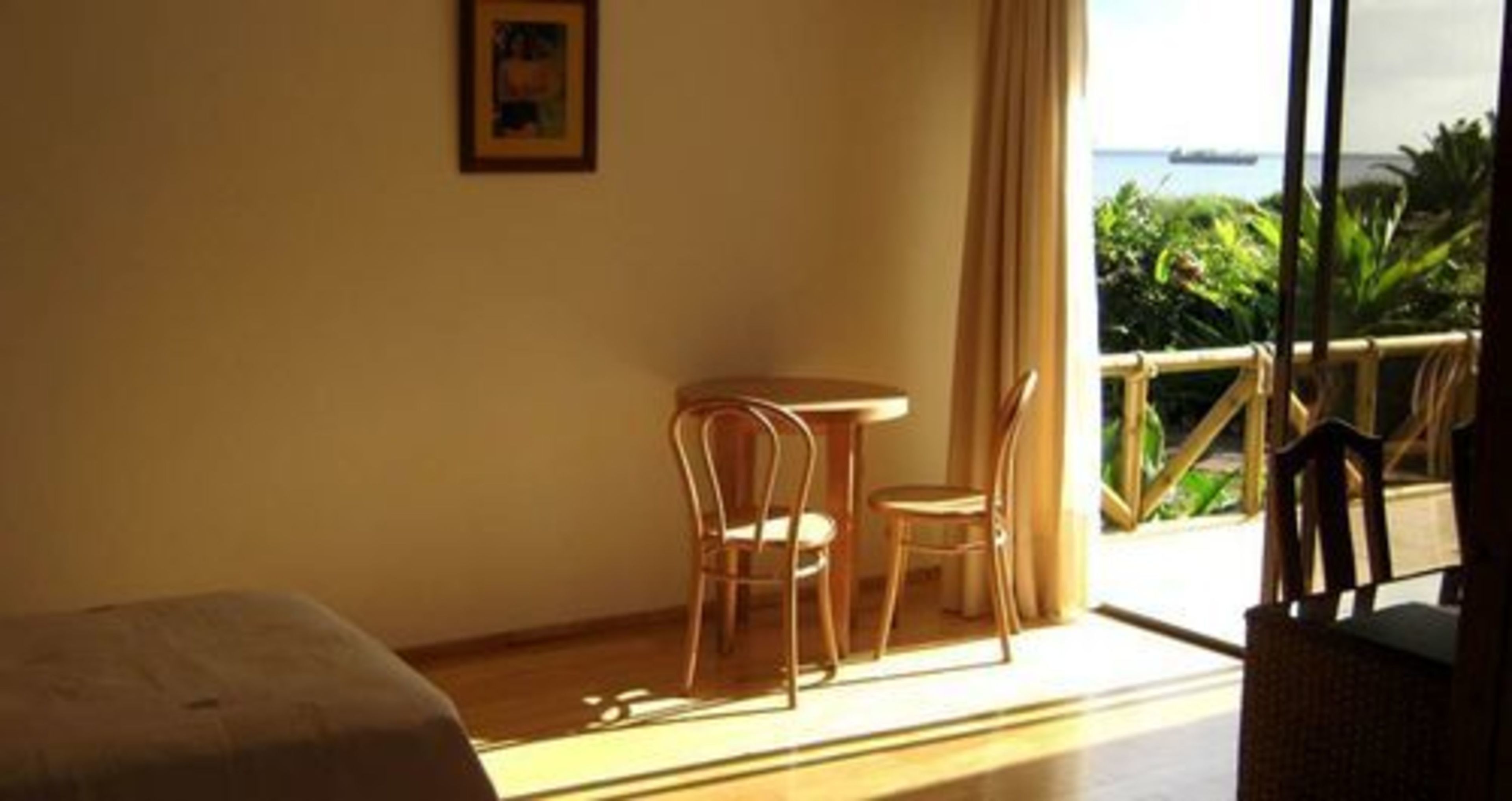 Een kamer met een bed, een klein rond tafeltje, twee stoelen en een groot raam met uitzicht op groen en een wateroppervlak.