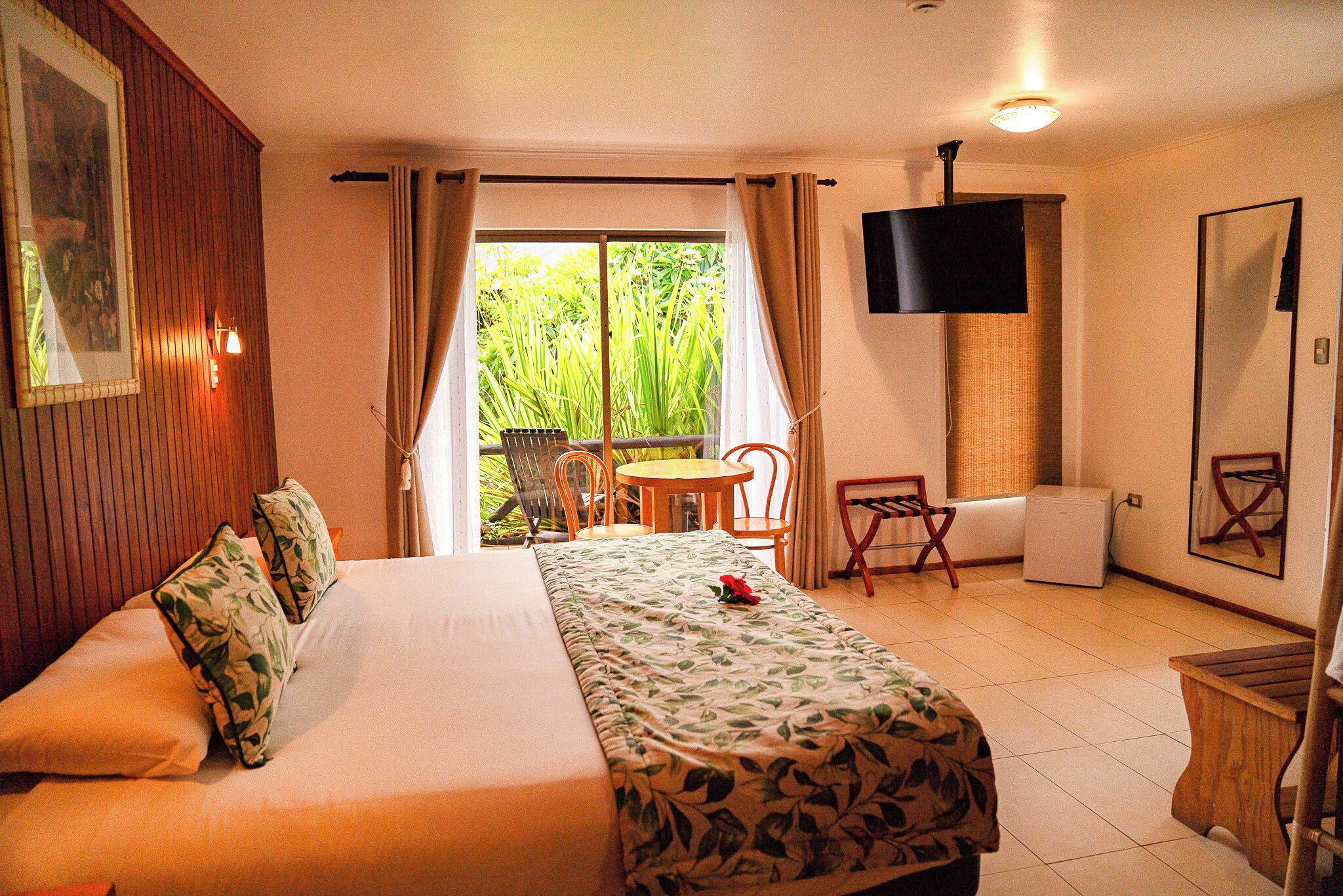 Habitación de hotel con cama, una mesita, sillas, televisión y balcón con plantas.
