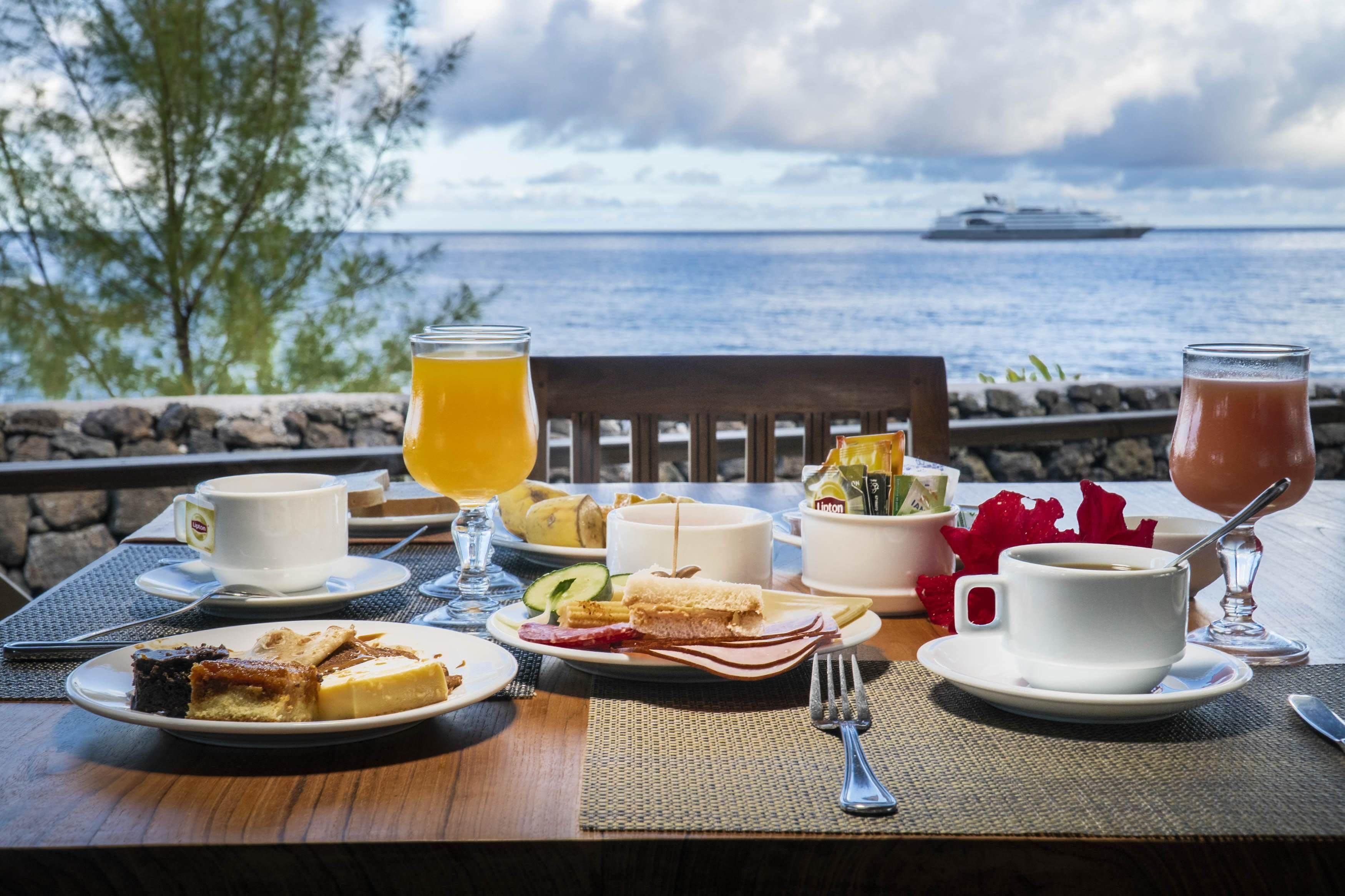 A table set with breakfast items including a glass of orange juice, a cup of coffee, and various pastries, with a scenic ocean view in the background.