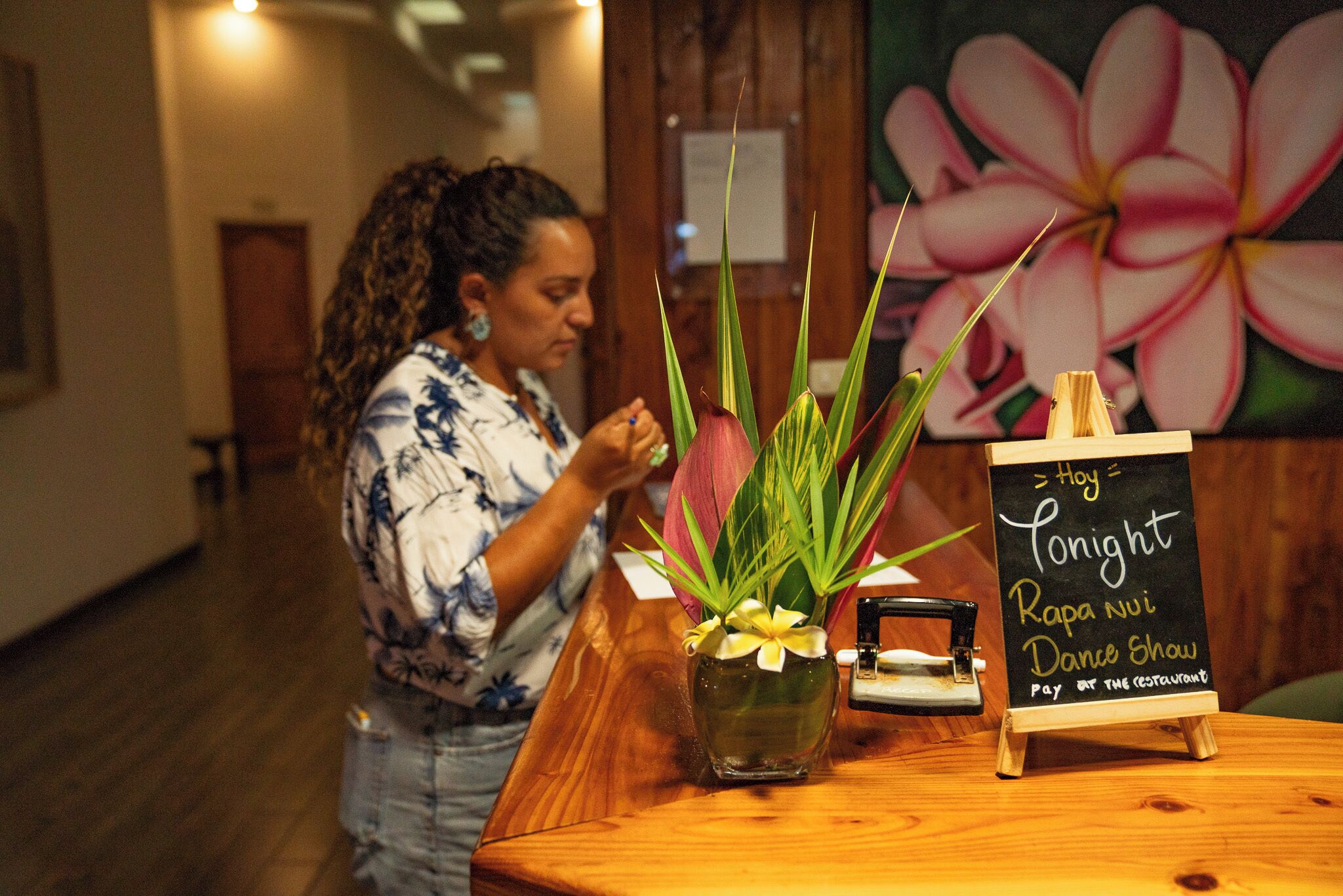 A woman at a wooden reception desk with a vase of flowers and a chalkboard sign.