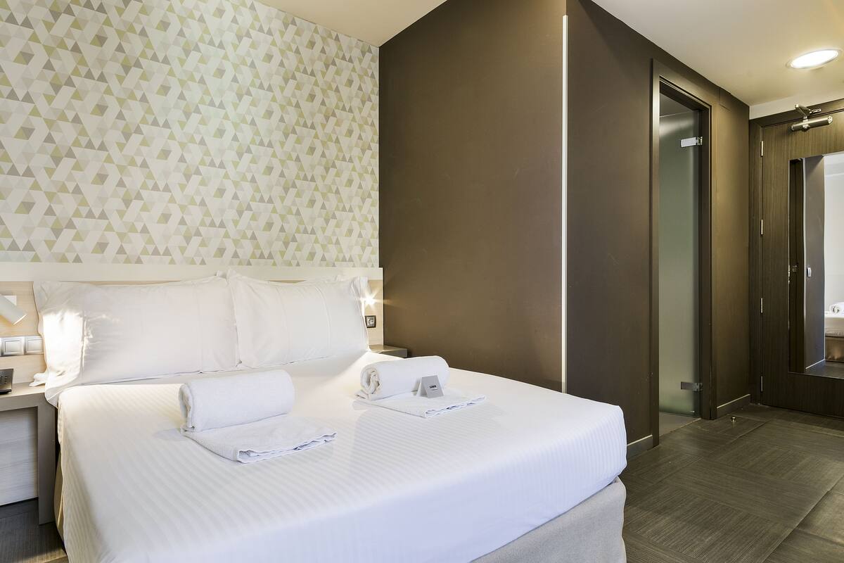 A hotel room with a neatly made bed, a nightstand with a lamp, and a modern geometric-patterned wall.