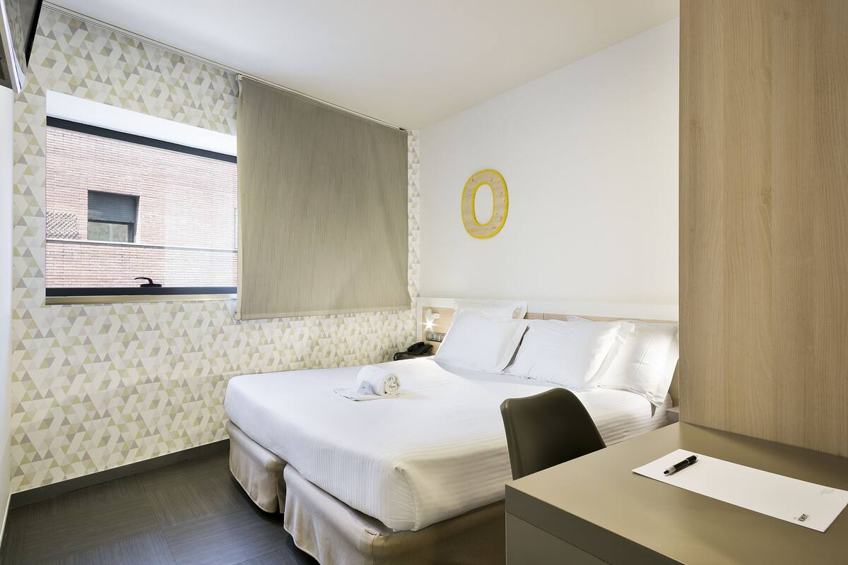 A hotel room with a bed, a desk, and a window with a view of a building.