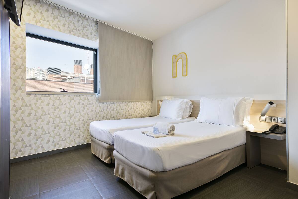 A hotel room with two beds, a window with a view of buildings, a wall-mounted letter 'n', and a bedside table with a lamp and phone.