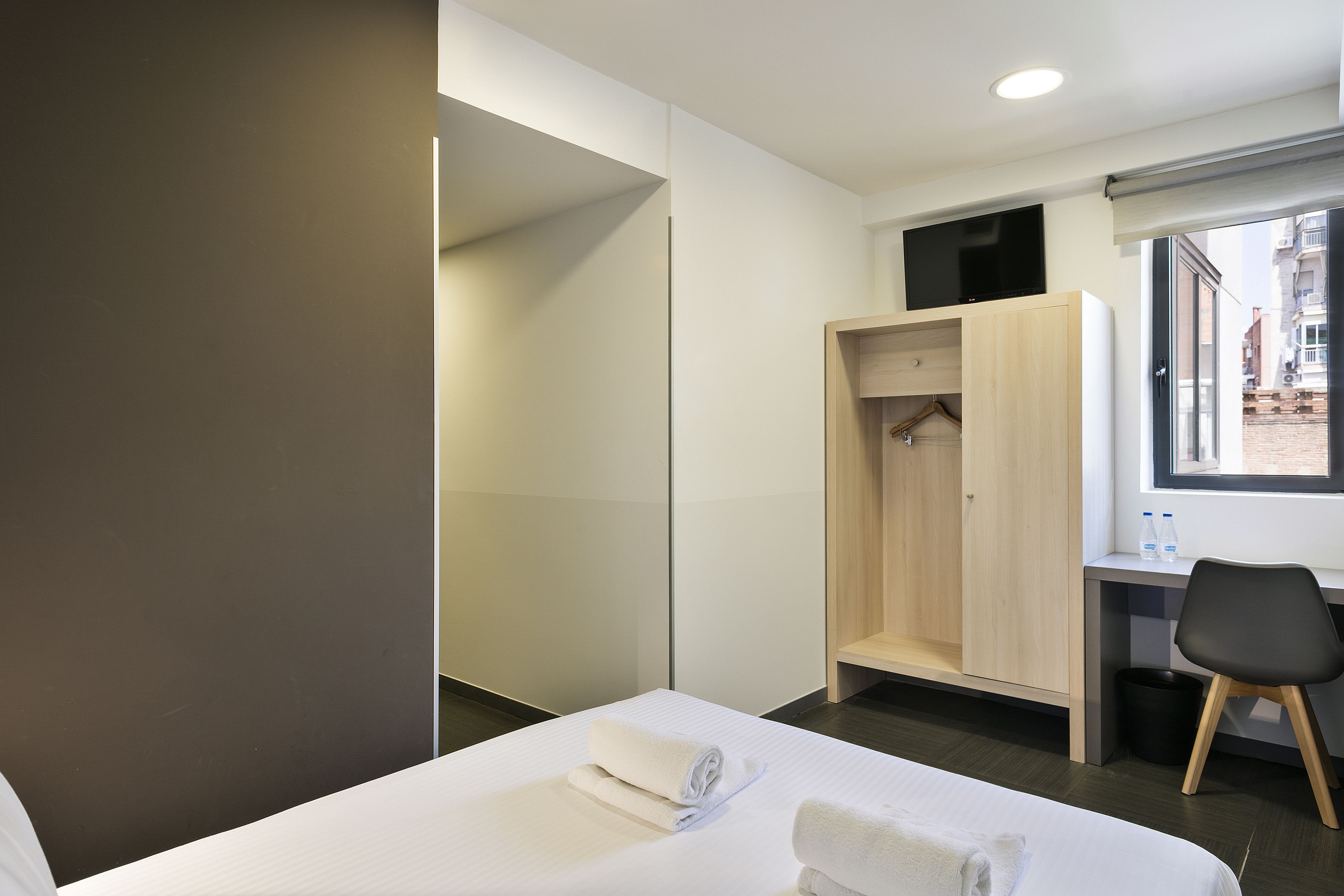 A modern hotel room with a bed, a desk with a chair, a wardrobe, and a TV on the wall.
