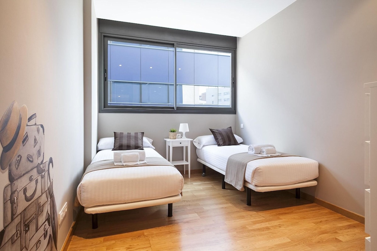 Habitación de hotel pequeña y moderna con dos camas individuales, una mesita de noche con lámpara y un ventanal con vistas a la ciudad.