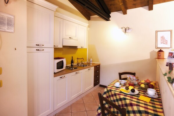 Una cocina compacta con armarios blancos, un microondas y un lavamanos. Hay una mesa de comedor con mantel a cuadros y una silla.