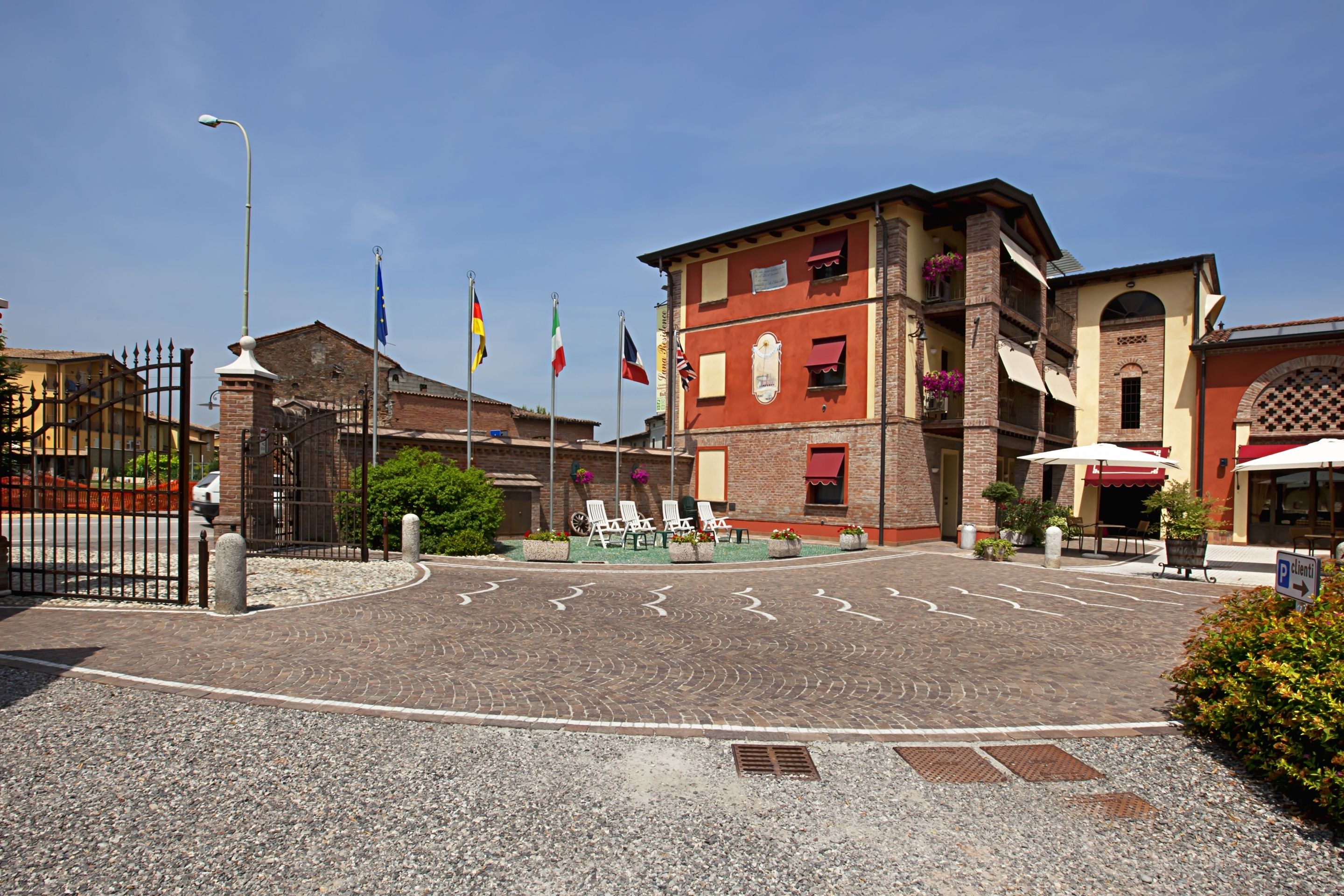 Un patio con zona pavimentada, mobiliario exterior y un edificio con varias banderas.