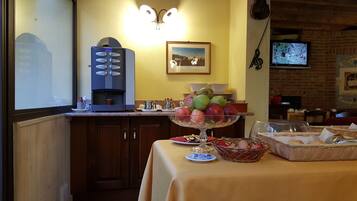 A breakfast setup with a coffee machine, fruit bowl, and breakfast items on a table.