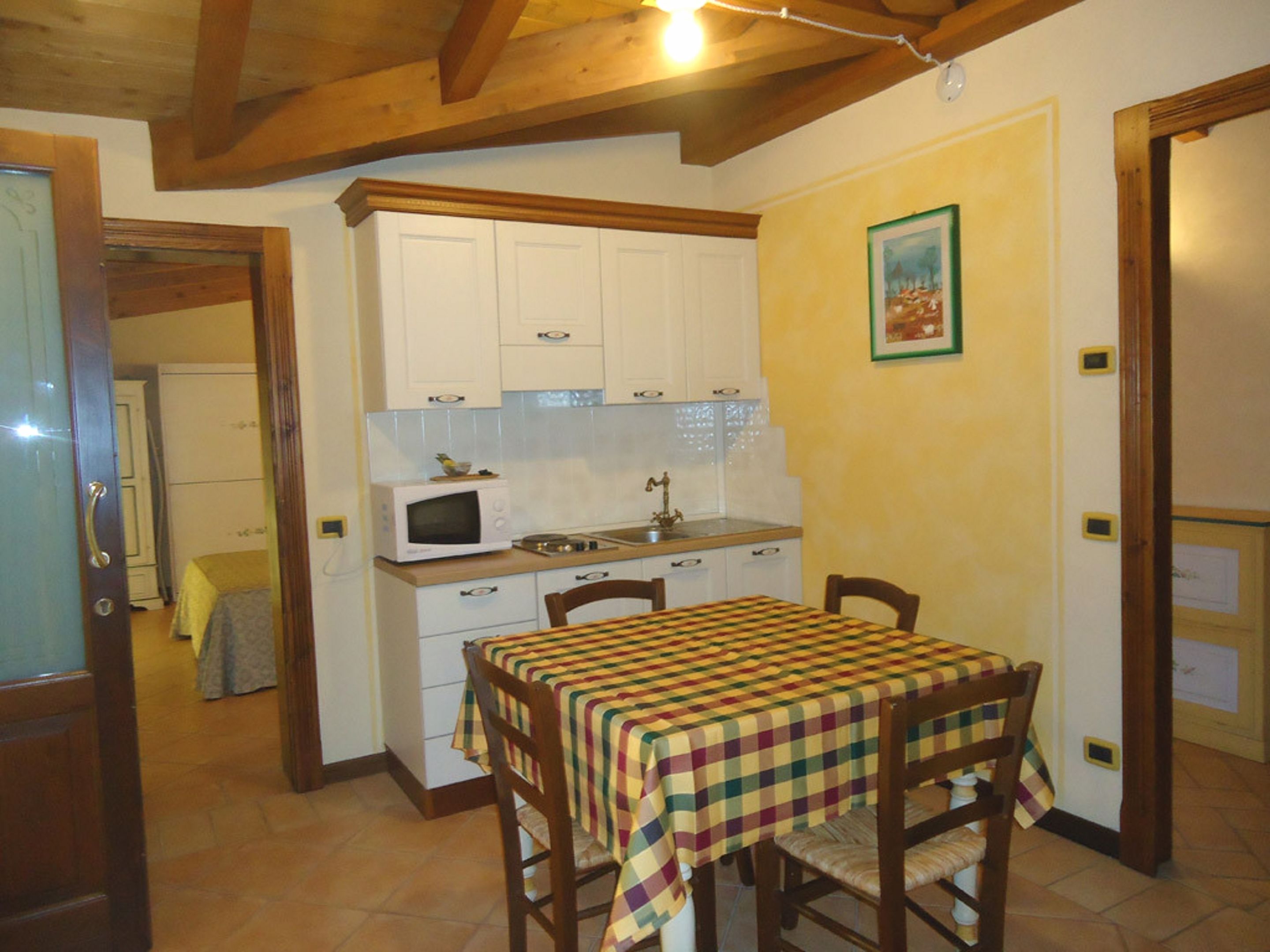 A compact kitchen with white cabinets, a microwave, and a stove. There is a table with a checkered tablecloth and wooden chairs.