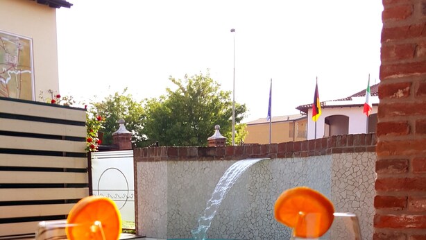 Dos vasos de bebida roja con rodajas de naranja sobre una mesa de madera, un ramo de rosas rojas, y una vista de una piscina con cascada al fondo.