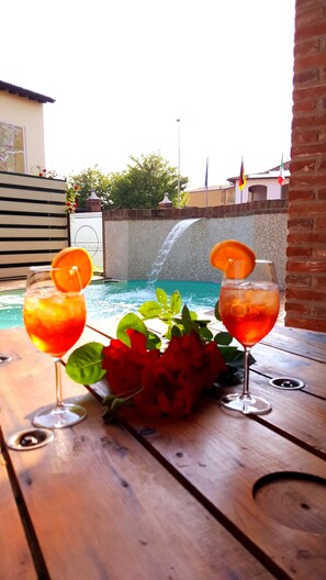 Dos vasos de bebida roja con rodajas de naranja sobre una mesa de madera, un ramo de rosas rojas, y una vista de una piscina con cascada al fondo.
