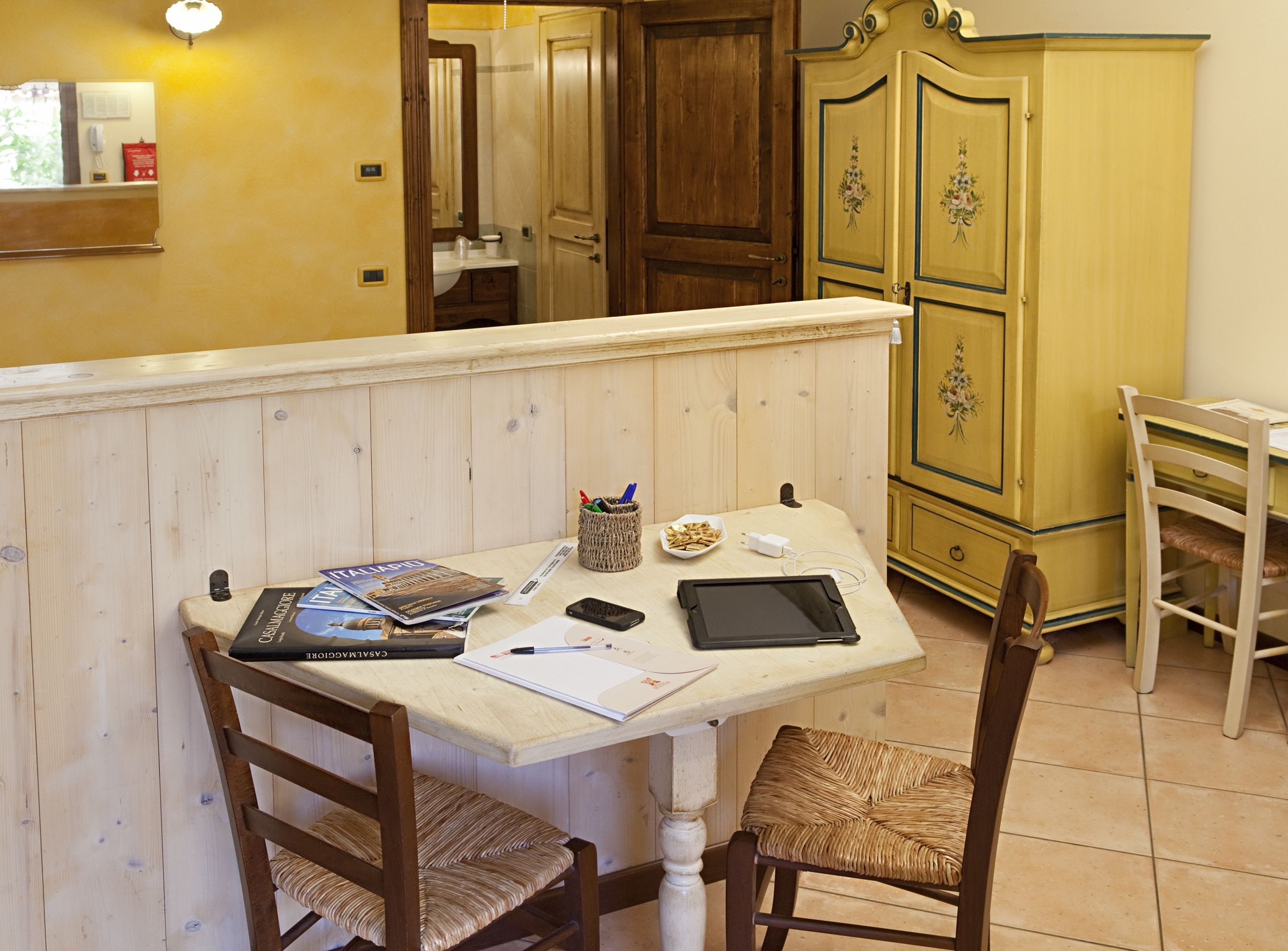 Ein Holztisch mit einem Laptop, einem Smartphone, einem Tablet und einigen Papieren. Es gibt Korbstühle und eine Kommode mit floralen Verzierungen.