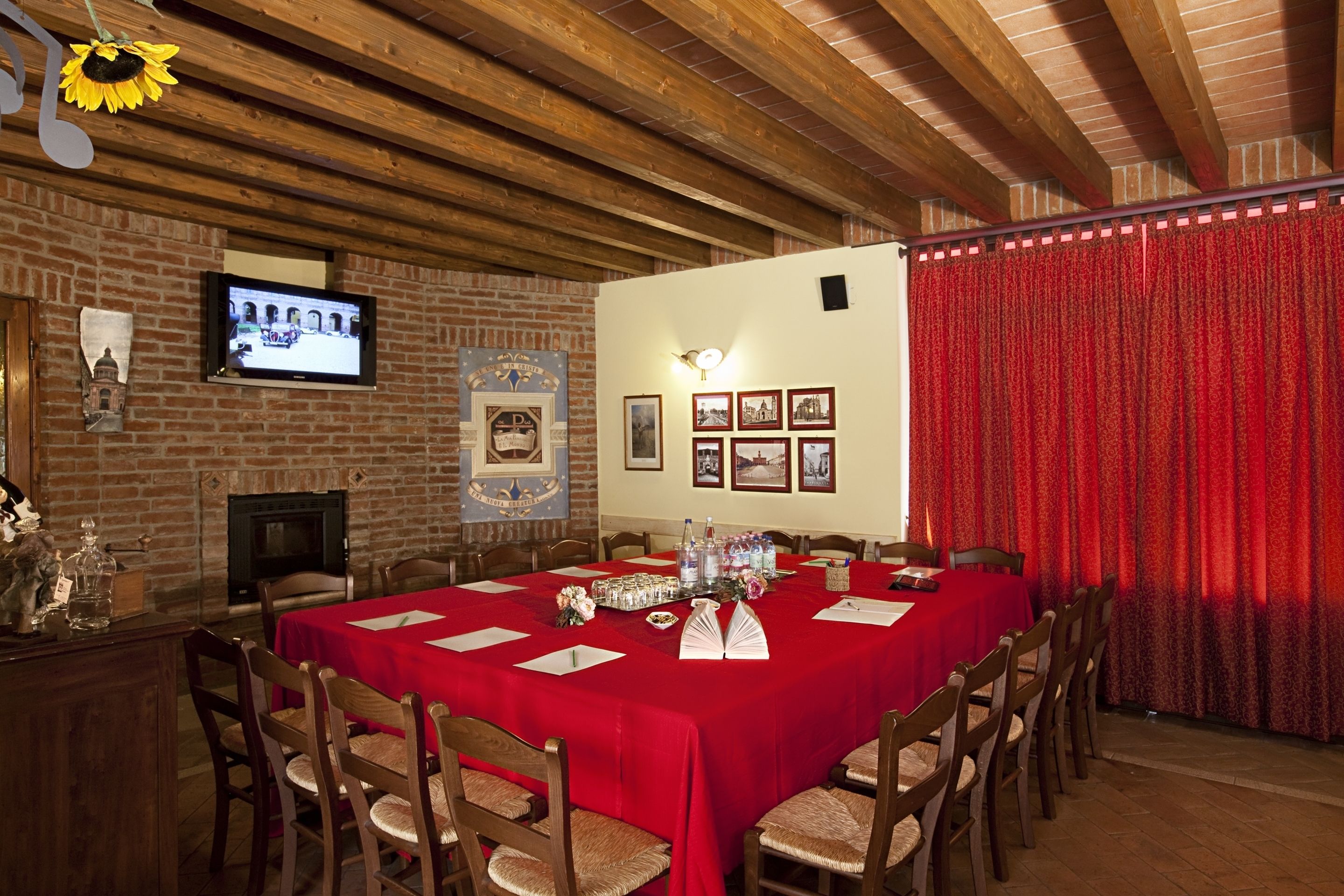 Un'area pranzo con un tavolo lungo ricoperto da una tovaglia rossa, sedie in legno, un muro in mattoni, un camino e un televisore montato a parete.