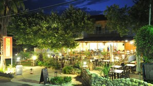 Un área de comedor al aire libre con paredes de piedra y mesas, rodeada de vegetación y un edificio con porche cubierto.