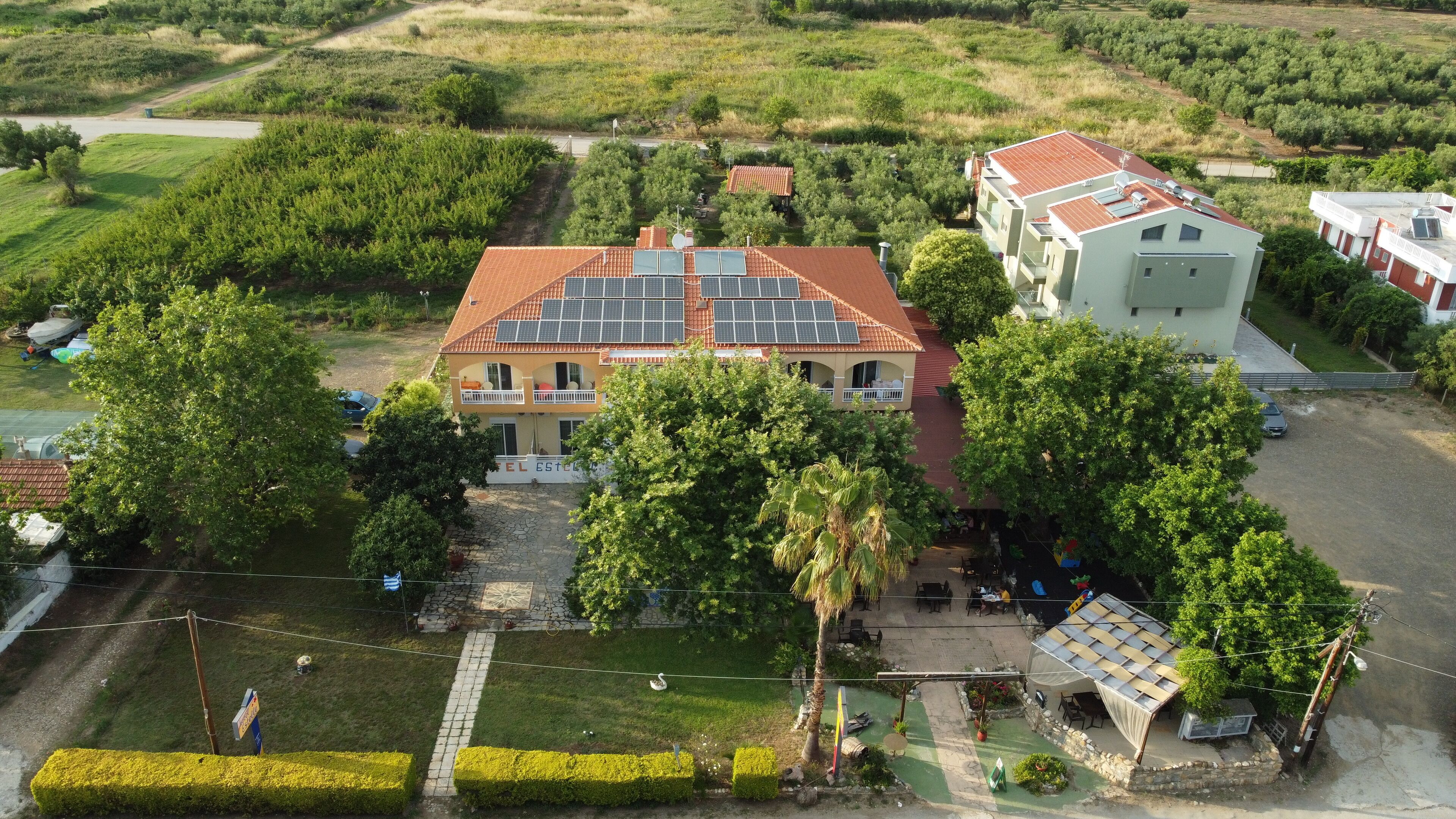 An aerial view of a hotel with a red-tiled roof, solar panels, and a garden area with a palm tree.