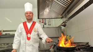 Un chef con uniforme blanco y una medalla, cocinando en una sartén sobre una hornalla en una cocina.