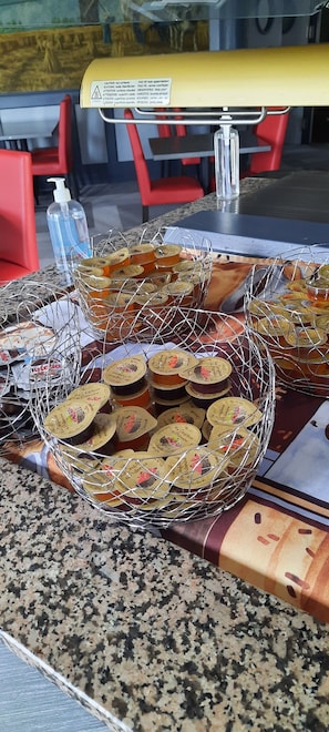 Una mesa de buffet con una cesta metálica que contiene frascos de comida, un dispensador de gel hidroalcohólico y un mantel amarillo.