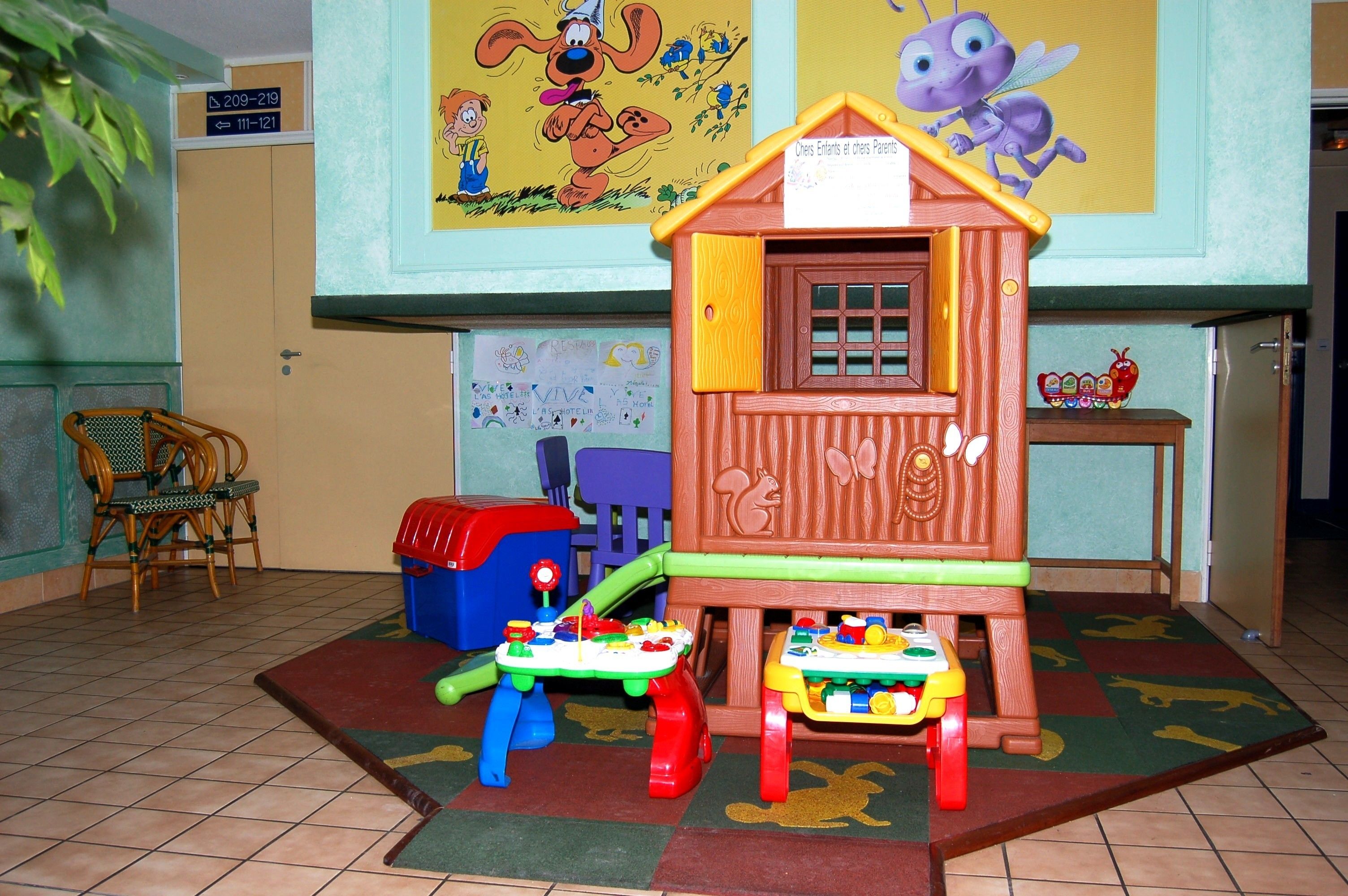 A children's play area with a wooden playhouse, a table with toys, and a mural on the wall.