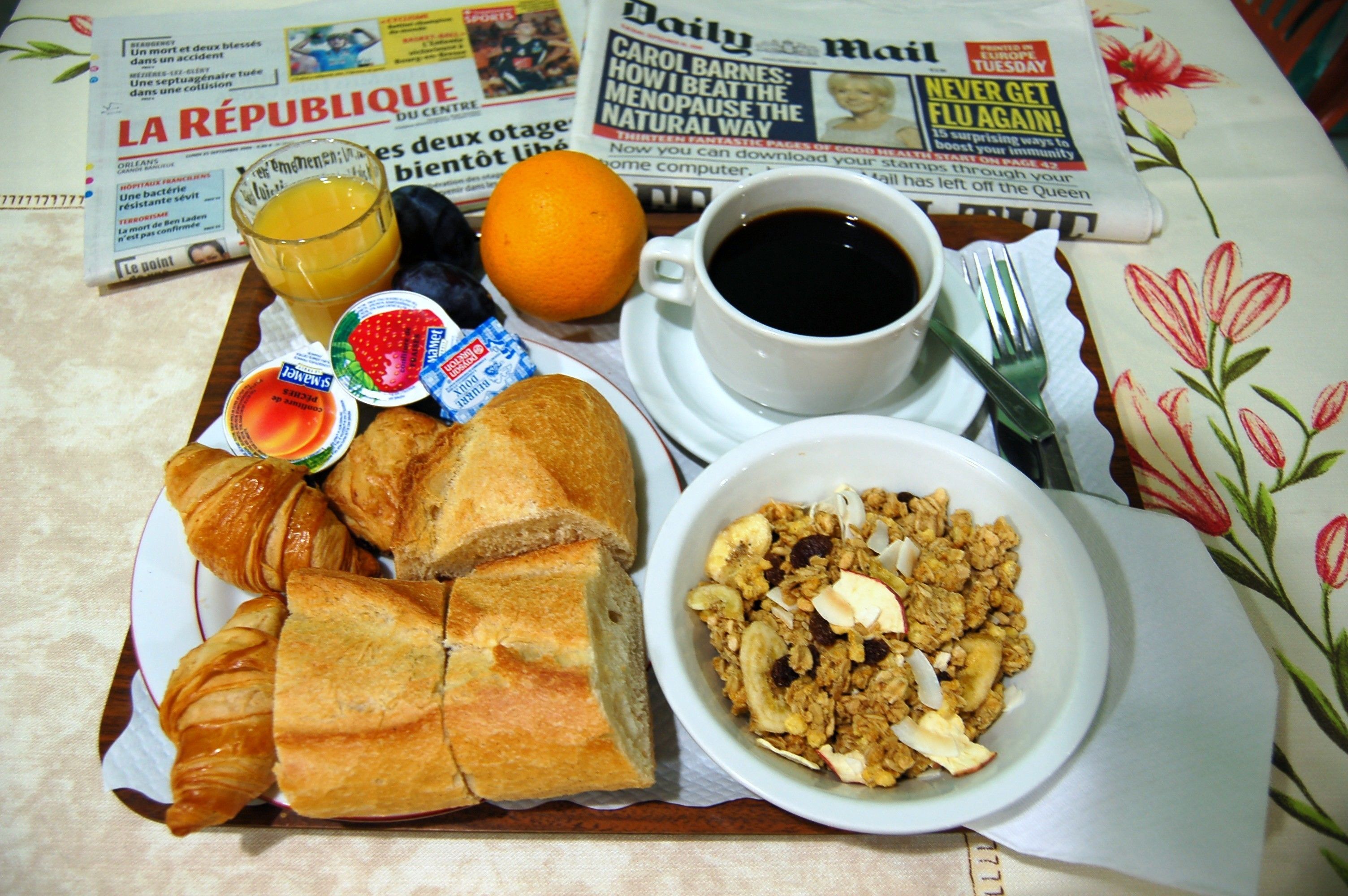 A breakfast tray with croissants, a bowl of granola, a glass of orange juice, and a cup of coffee, placed on a table with newspapers.