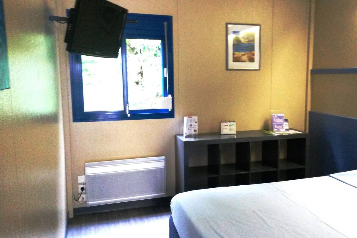 Habitación de hotel con cama, ventana, estante con objetos y televisor en la pared.