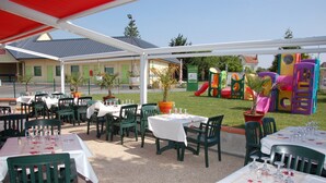 Un patio al aire libre con mesas y sillas, un área de juegos infantiles al fondo y un edificio con una puerta verde.