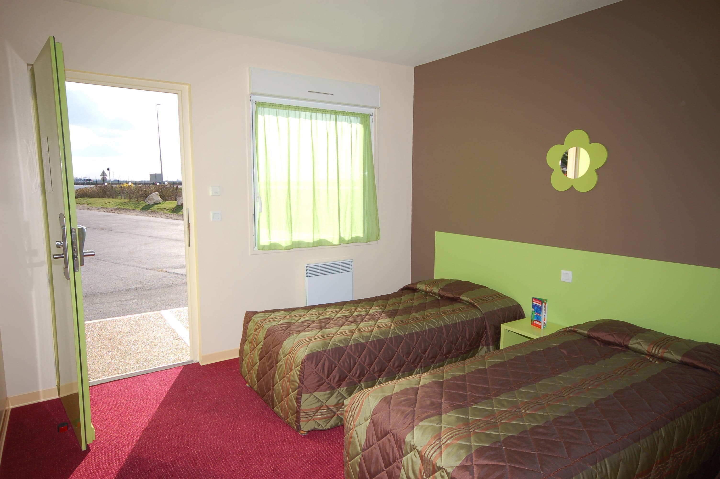 A room with two beds, a window with curtains, a door, and a small table with a book.