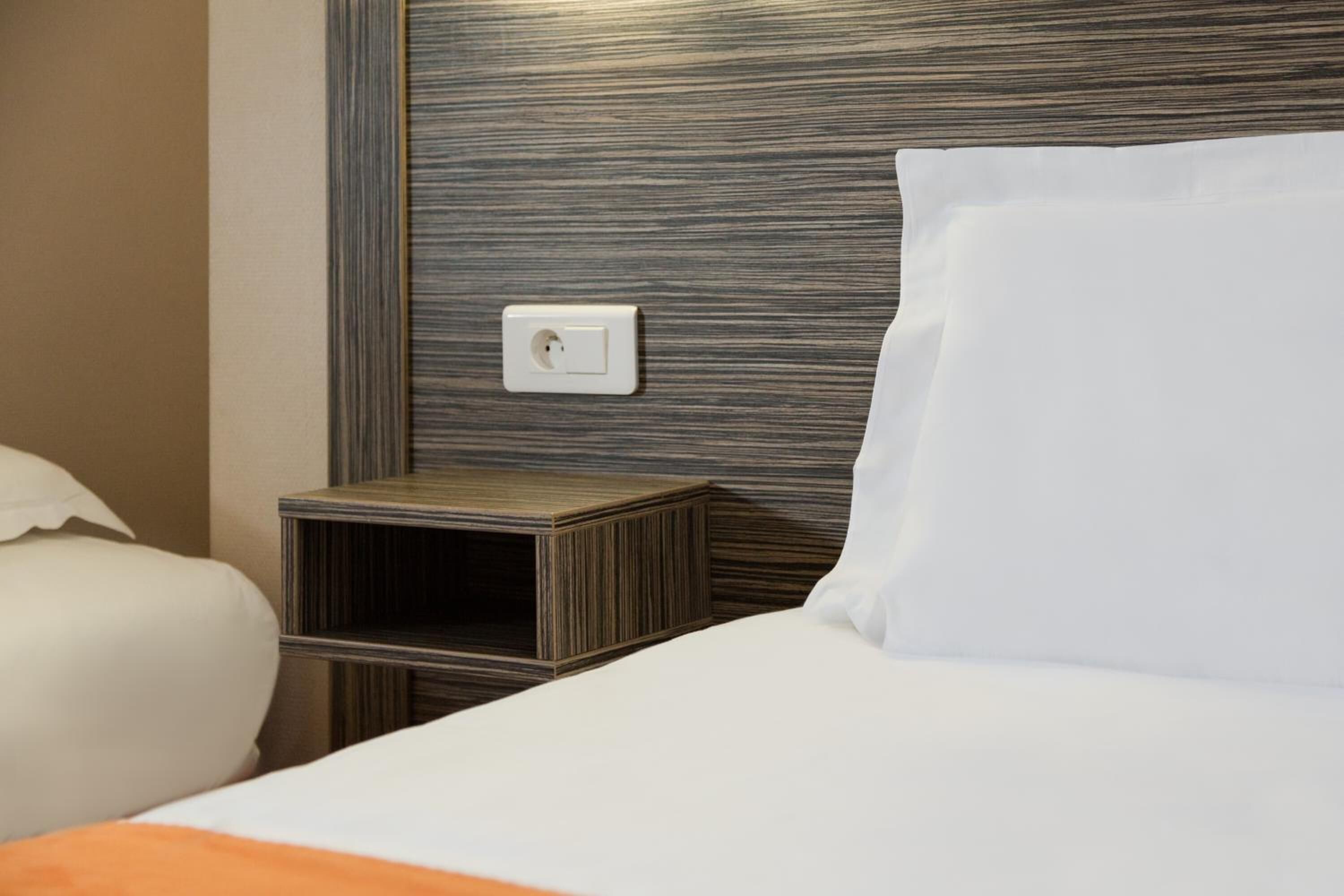 Una habitación de hotel con una cama bien hecha, una mesita de noche de madera y un enchufe en la pared.