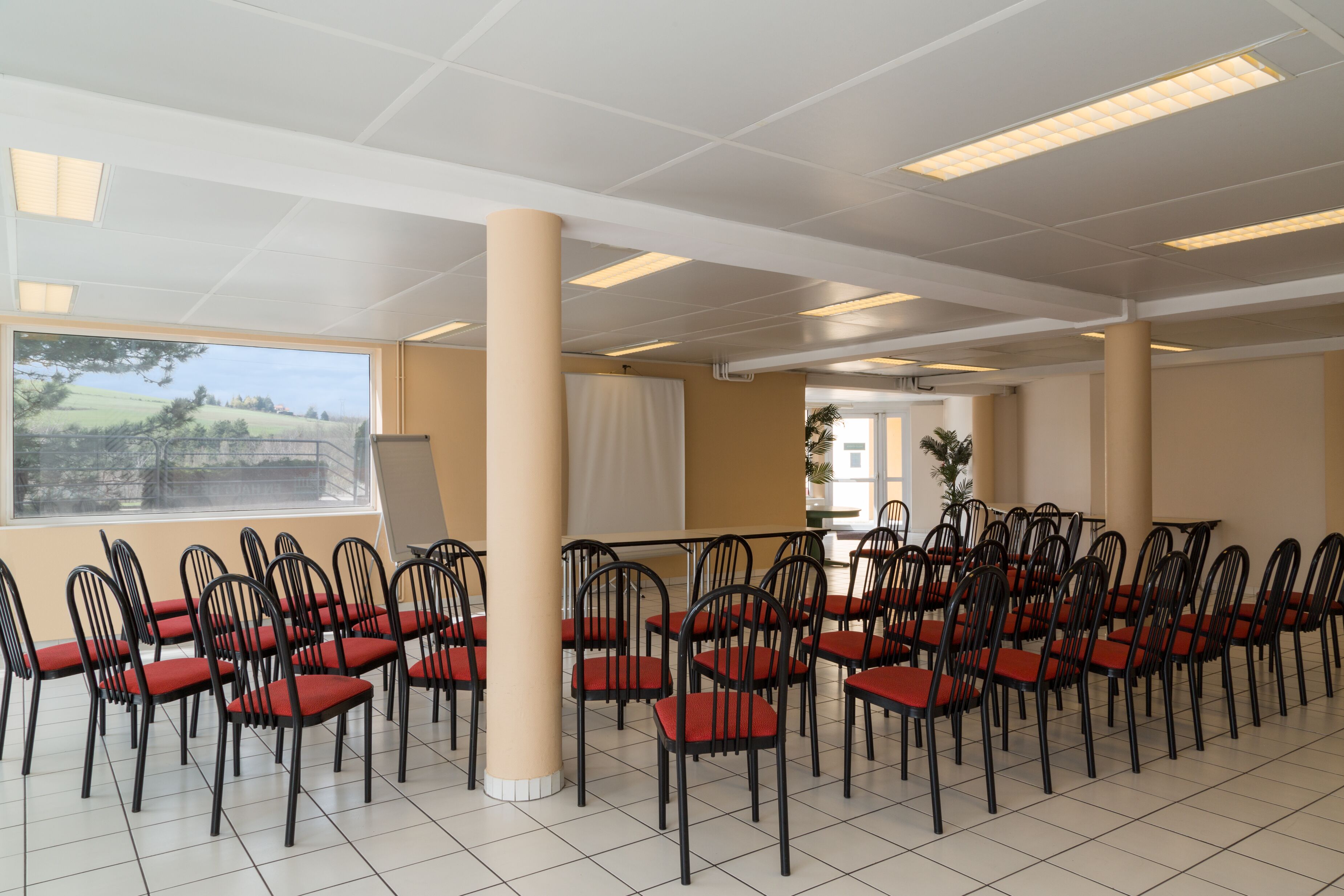 Une salle de conférence avec des rangées de chaises noires dotées de coussins rouges, des murs de couleur beige et de grandes fenêtres donnant sur un paysage verdoyant.