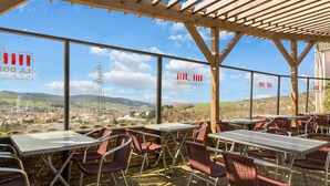 An outdoor café with tables and chairs, large windows offering a view of a town and hills, and a wooden pergola structure.
