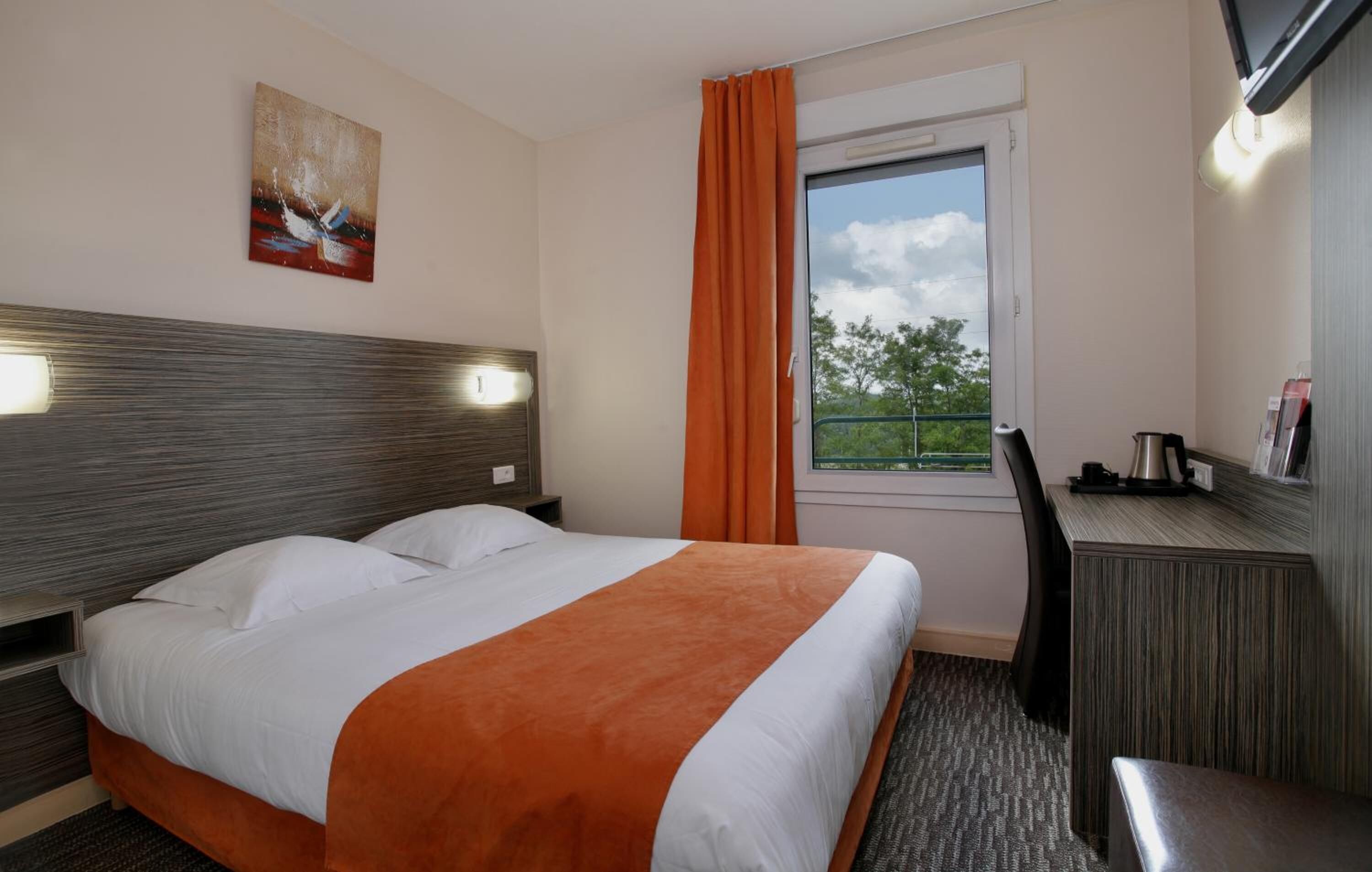 Une chambre d’hôtel avec un lit, un bureau, une chaise, une fenêtre avec des rideaux, un tableau au mur et une vue sur les arbres à l’extérieur.
