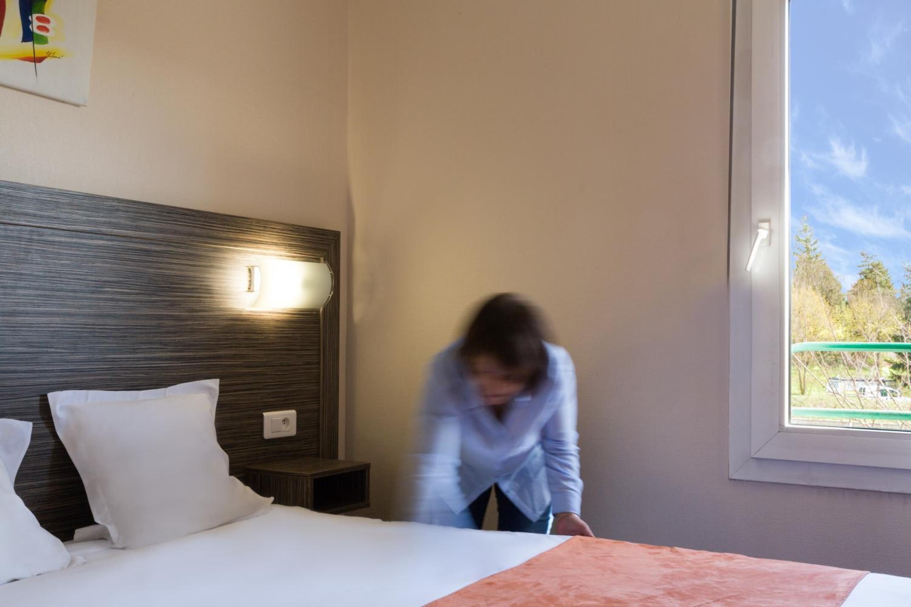 A hotel room with a bed, a nightstand, a wall-mounted light, and a window with a view of trees.