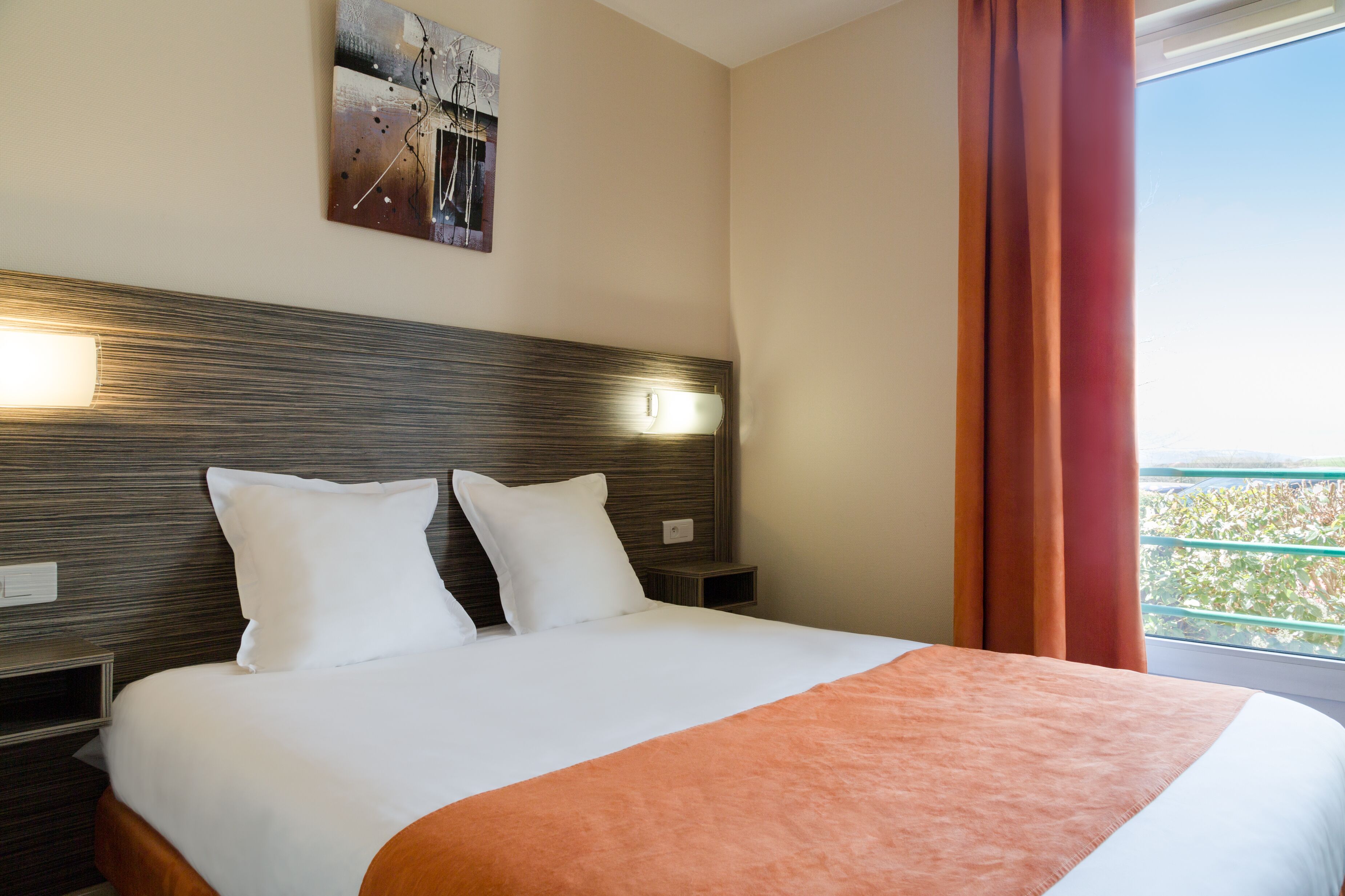 A hotel room with a large bed, two bedside tables, a headboard with a light fixture, a painting on the wall, and a view of greenery through the window.