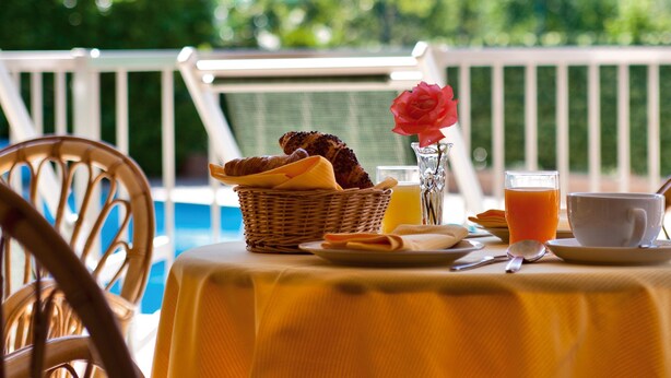 Mesa posta com itens de café da manhã, um vaso com uma flor e um copo de suco de laranja numa varanda com vista para as montanhas.
