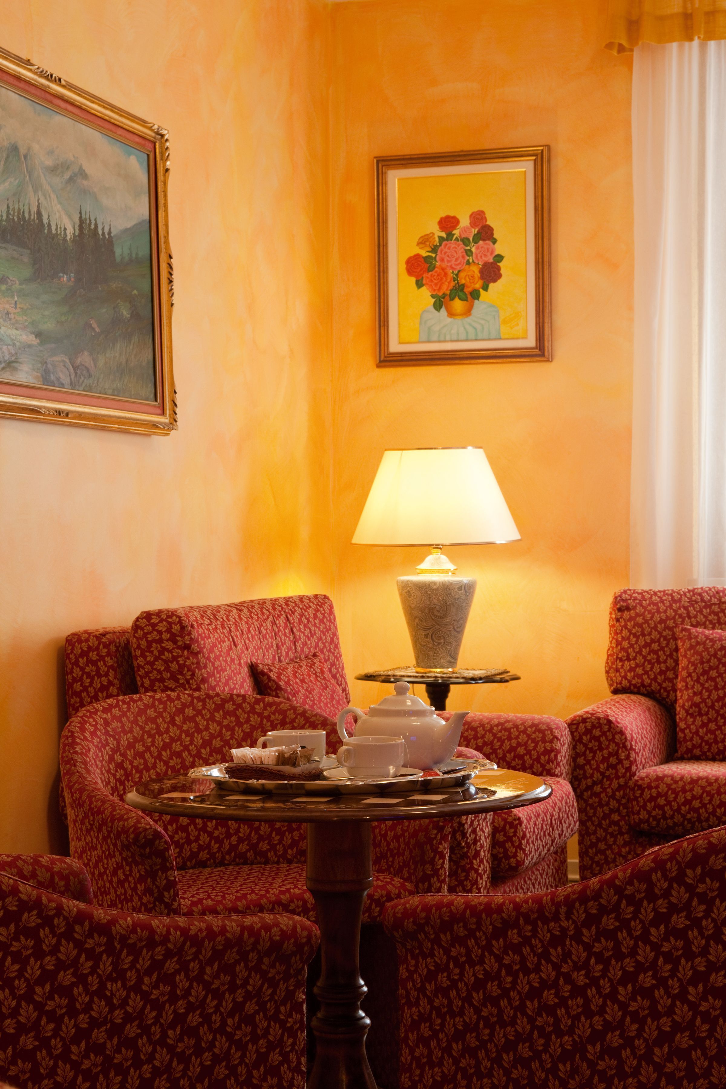 A hotel room with red patterned sofas, a small round table with a teapot and cups, a lamp, and two framed artworks on the walls.
