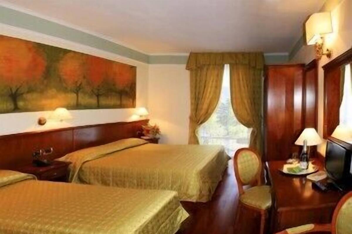 A hotel room with two beds, a desk with a chair, a painting on the wall, and a window with curtains.