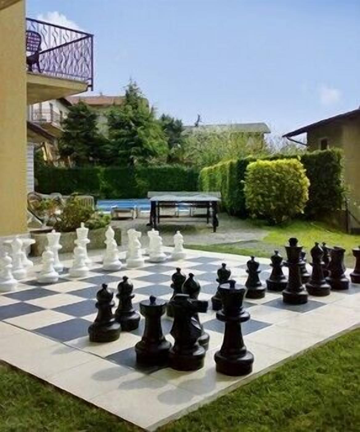 A large outdoor chessboard set up on a patio with a table and chairs in the background.