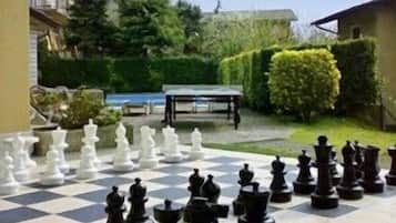 A large outdoor chessboard set up on a patio with a table and chairs in the background.