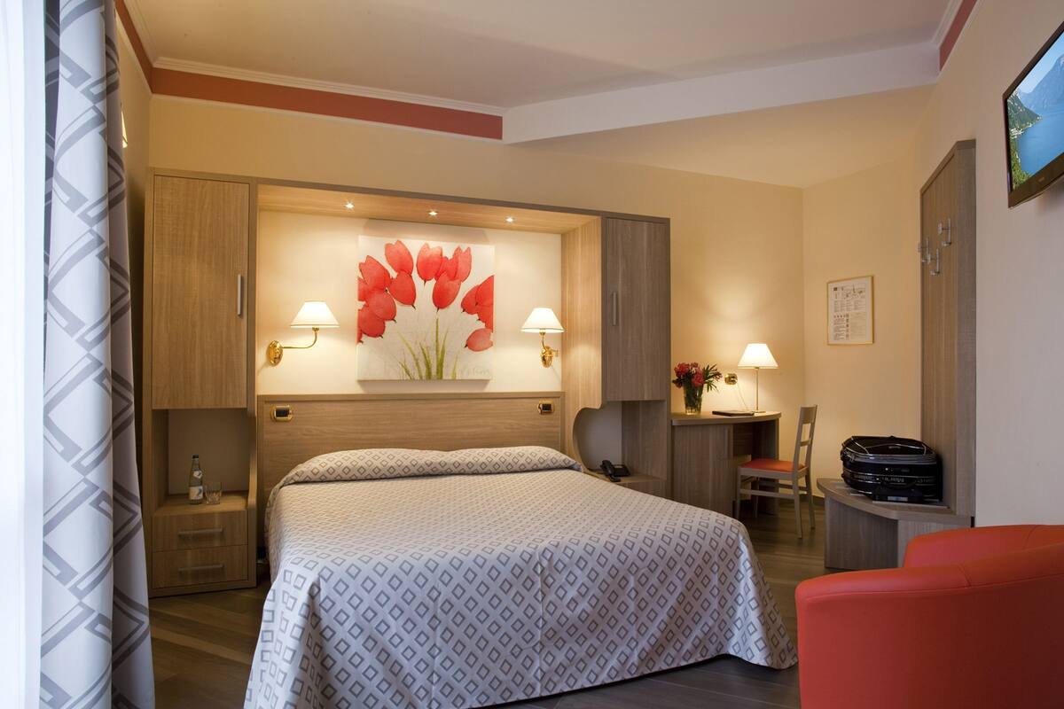 A hotel room with a large bed, two bedside tables, a desk with a chair, a red armchair, and a television on the wall.