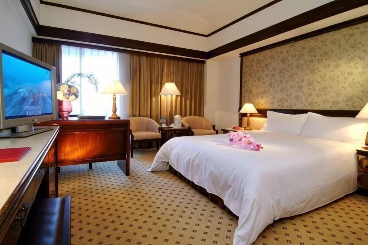 A hotel room with a bed, a desk, a television, two lamps, and a window with curtains.
