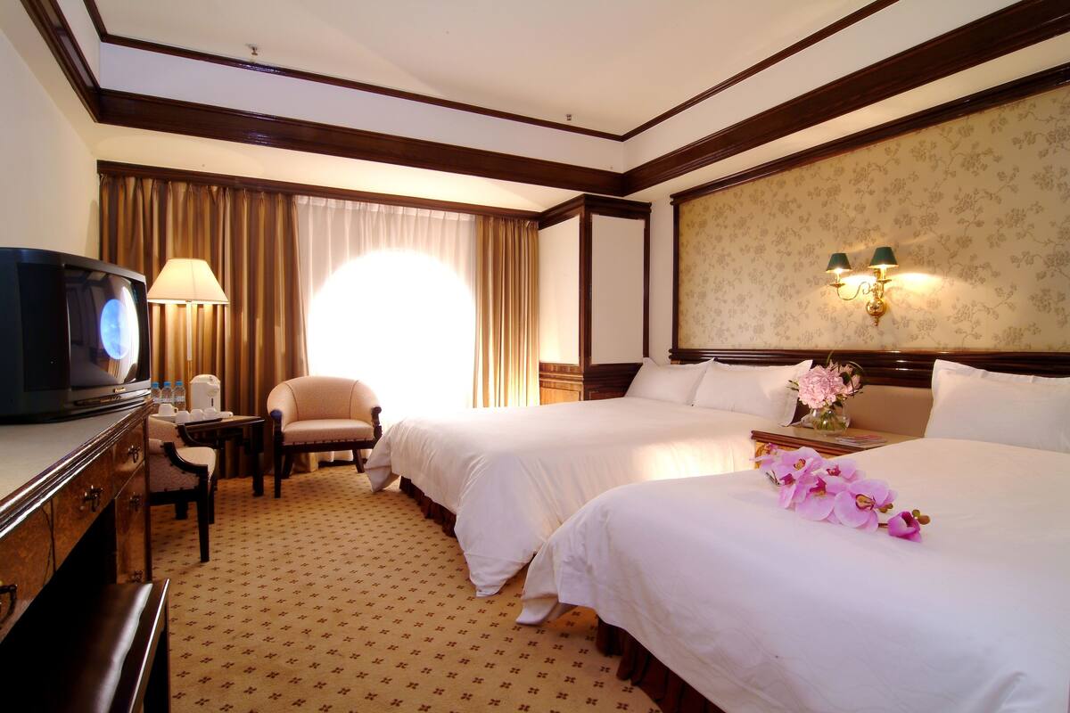 A hotel room with two beds, a desk with a television, a chair, and a lamp.