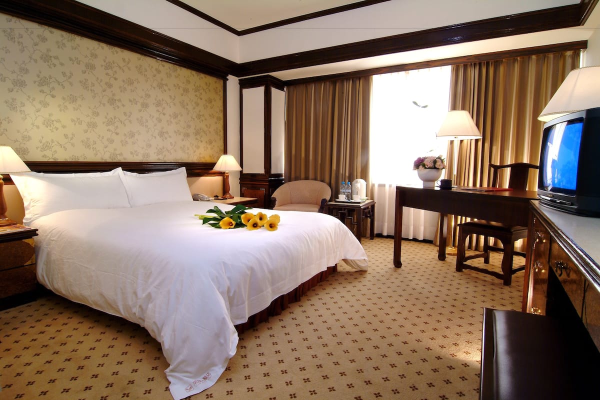 A hotel room with a large bed, two bedside tables, a desk with a chair, a television, and a window with curtains.