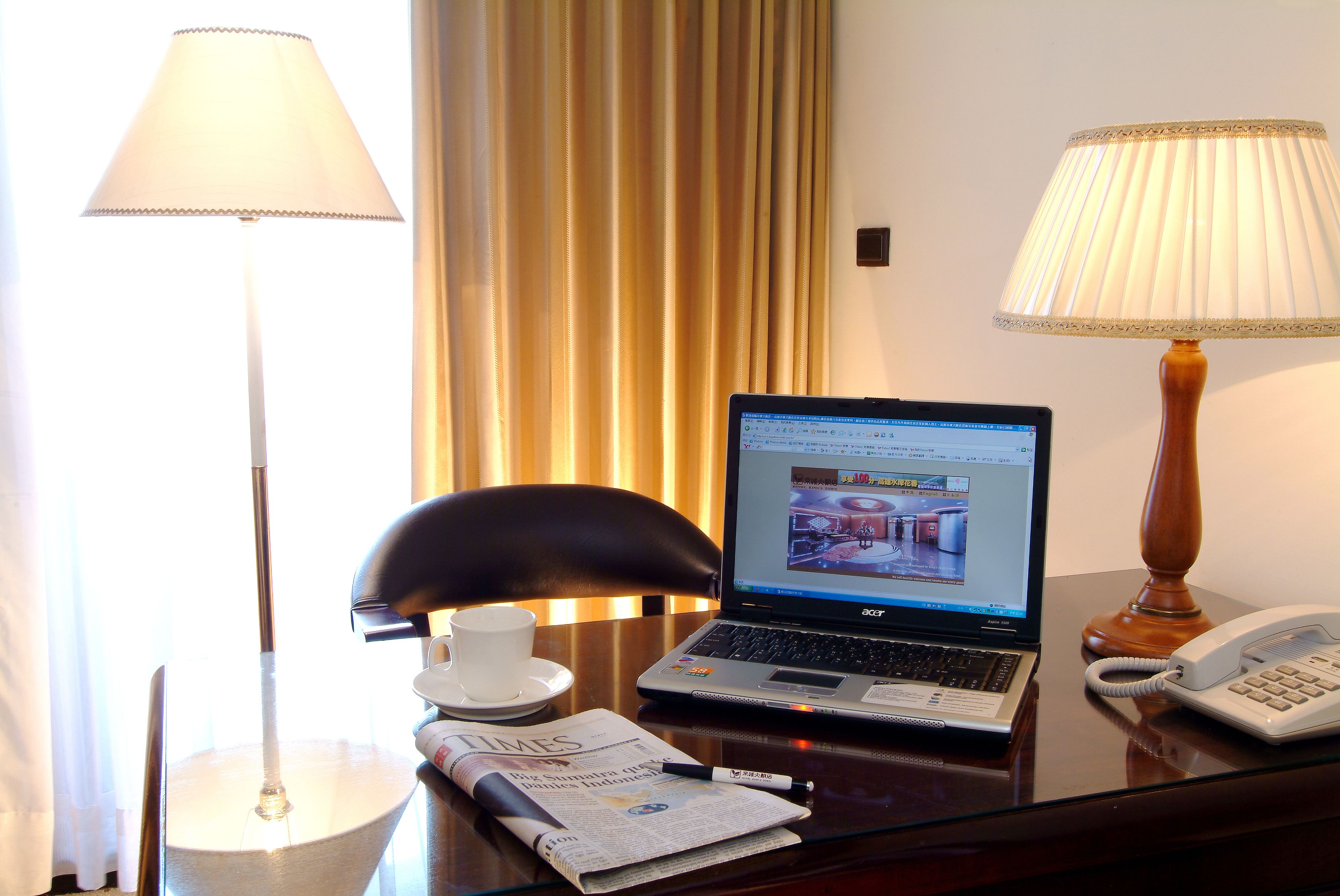 A hotel room desk with a laptop, a telephone, a lamp, a chair, a cup, and a newspaper.